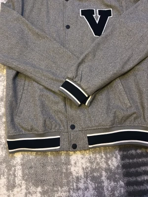 Grå bomberjacka med V från Dressmann - Snygg grå bomberjacka från Dressmann med svart och vit ribbade muddar och krage. Jackan har tryckknappar och är gjord i mjukt material, perfekt för de som gillar och  ha översize och en avslappnad streetstil priset kan diskuteras !!!!!!!!!!