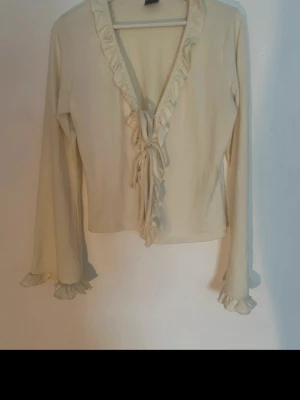 Krämvit volangkofta med knytband - Söt krämvit kofta med volangkant längs urringningen och knytband framtill. Långa ärmar med volangdetalj vid ärmslut. Perfekt för lager-på-lager och en romantisk look. Storlek L