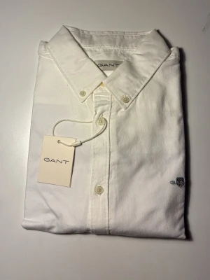 Vit skjorta från GANT med knappar - Klassisk vit skjorta från GANT med button-down krage och broderad logga på bröstet. Skjortan har knappar framtill och är tillverkad i mjuk bomull. Perfekt för en clean och stilren look. Köpte de för 800 kr och den är för liten för mig därför har jag bestämt mig att sälja den. Den är M fast passar även Large