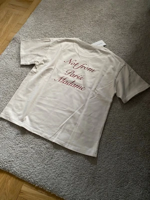 Drole De Monsieur Not From Paris Madame Tshirt - Säljer en vit t-shirt Not From Paris Madame med tryck i röd text både fram och bak. T-shirten har klassisk passform och korta ärmar. Perfekt för dig som gillar stilrena plagg med en twist.