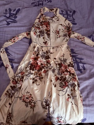 Blommig halterneck jumpsuit - Säljer en superfin blommig lång jumpsuit med halterneck och öppen rygg. Den har ett krämfärgat tyg med rosa, röda och gröna blommor, samt knytband i midjan och nacken. Perfekt för dig som vill ha en luftig och somrig look. Materialet känns lätt och mjukt.