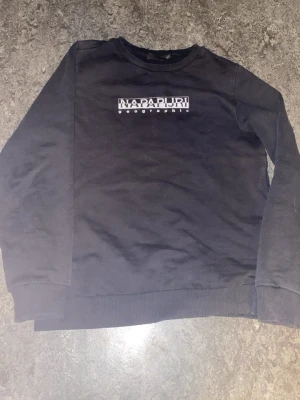 Svart sweatshirt från Napapijri - Svart sweatshirt från Napapijri med vit logotyp på bröstet. Tröjan har rund halsringning, ribbade muddar vid ärmslut och nederkant samt en klassisk passform. Perfekt för en avslappnad och stilren look.