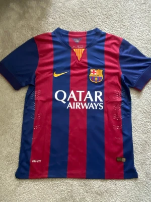 FC Barcelona Messi matchtröja - Säljer en klassisk FC Barcelona matchtröja från Nike med Messi 10 på ryggen. Tröjan har blå och röda vertikala ränder, klubbmärke och Qatar Airways-logga framtill samt Dri-FIT-material som andas. Perfekt för fotbollsfans och samlare!