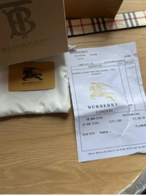 Rutig Burberry bälte i skinn - Säljer ett klassiskt Burberry bälte i skinn med det ikoniska rutiga mönstret i beige, svart, rött och vitt. Bältet har ett elegant spänne i guld och kommer med originalpåse och box. Perfekt accessoar för att lyfta din outfit.