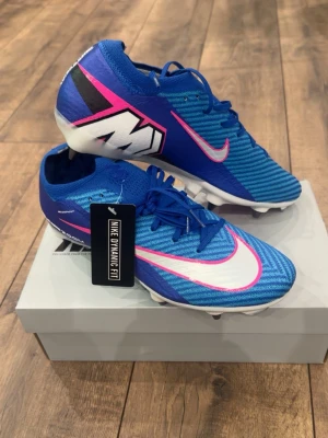 Nike blåa Mercurial fotbollsskor - Snygga blåa Nike Mercurial fotbollsskor med vita och rosa detaljer samt stor vit Nike-logga på sidan. Skorna har snörning, låg profil och taggar för bra grepp på planen. Tillverkade i syntetmaterial med Nike Dynamic Fit för optimal passform.