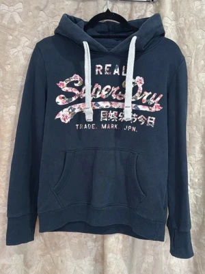 Fet Superdry hoodie - Mör resident iconic hoodie från Superdry med stor magficka, vit dragsko och blommigt tryck på bröstet. Klassisk huva och mjukt material, ver riktigt chill och passar perfekt till jeans eller leggings. Snygg logga och japanska tecken framtill.