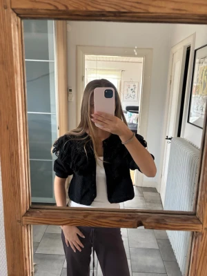 Svart kort kofta med volanger - Trendig svart kofta med kort passform och stora volangdetaljer över axlarna. Koftan har knappar framtill och puffiga ärmar som ger en cool och unik look. Perfekt att styla över ett linne eller t-shirt för en extra touch.