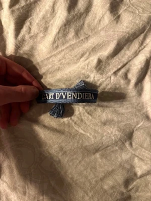 Blått tygarmband L'ART D'VENDIERA - Snyggt blått tygarmband med texten 'L'ART D'VENDIERA' broderat i vitt. Armbandet har dekorativa tofsar i ändarna och justerbar knytning. Perfekt accessoar för en personlig touch till din stil.