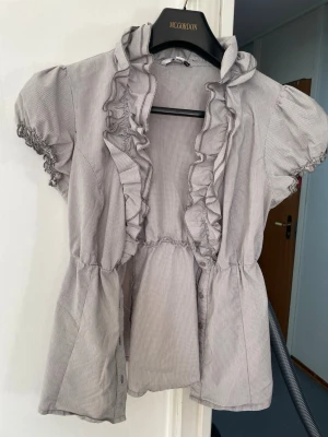 Gråblus med volang och puffärm - Söt gråblus från Tally Weijl med små rutor, volangkant längs knäppningen och puffiga korta ärmar med resår. Blusen har markerad midja och lätt utsvängd passform. Perfekt för dig som gillar romantiska detaljer och en feminin look.