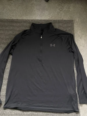 Svart träningströja från Under Armour - Säljer en svart långärmad träningströja från Under Armour med halv dragkedja framtill och hög krage. Tillverkad i ett lätt och stretchigt funktionsmaterial som passar perfekt för träning eller löpning. Diskret logga på bröstet.