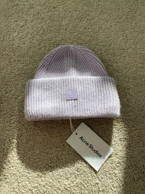 Acne Studios Ull Beanie Small Face Logo - Säljer en ljuslila ribbstickad mössa från Acne Studios med uppvikt kant och den klassiska Acne-loggan framtill. Mössan är gjord i mjukt material och har en stilren, minimalistisk look som passar perfekt till kalla dagar.