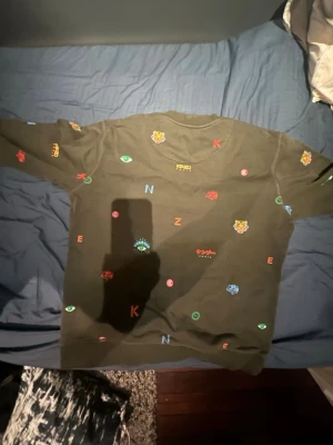Mörkgrön Kenzo sweatshirt med tryck - Säljer en mörkgrön sweatshirt från Kenzo med färgglada broderade och tryckta detaljer som tigrar, ögon och bokstäver. Tröjan har rund hals och är tillverkad i mjuk bomull. Perfekt för dig som gillar streetwear och unika prints.