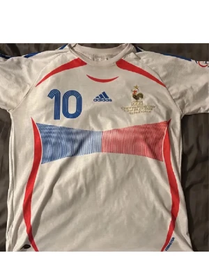  Zidane T - shirt  - Säljer en vit Frankrike t shirt från Adidas med blå och röda detaljer. Tröjan har korta ärmar och nummer 10 på framsidan samt namnet 'Zidane' på baksidan. Från final 2006 Italien vs Frankrike. LIMITED EDITION! Haft den bara hängd uppe i rummet. Först till kvarn!