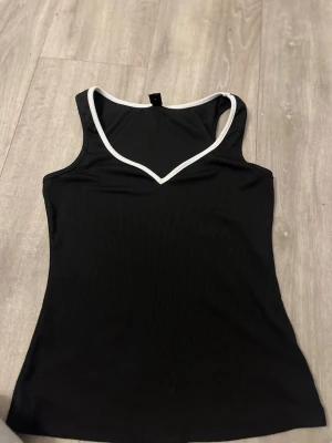 Svart ribbad linnetopp med vit kant - Snygg svart ribbad linnetopp med vit kant runt v-ringningen. Toppen är ärmlös och har en figurnära passform, perfekt att styla till jeans eller kjol. Enkel men trendig design som passar till många olika looks.