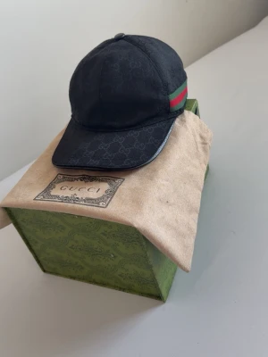 Gucci keps - Snygg svart keps från Gucci med diskret GG-monogram över hela kepsen och klassiska gröna och röda ränder på sidan. Kepsen har böjd skärm och justerbar rem bak. Tillverkad i canvasmaterial och levereras med originalkartong och dustbag.  Pris går att diskuteras, tar emot byten