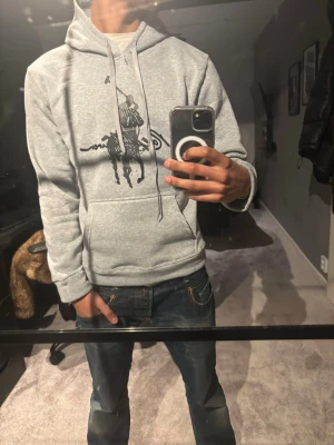 Grå hoodie med tryck från Ralph Lauren - Snygg grå hoodie från Ralph Lauren med klassiskt svart polospelar-tryck på bröstet. Hoodien har huva med dragsko och en stor magficka framtill. Perfekt för en avslappnad och trendig stil. Storlek M men passar som S