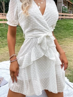 Vit klänning med volanger - Supersöt vit kortklänning med omlottmodell och knyt i midjan. Klänningen har volangdetaljer, prickigt mönster och korta puffärmar. Perfekt för sommarens alla tillfällen och ger en romantisk vibe. Tillverkad i ett lätt och luftigt material.