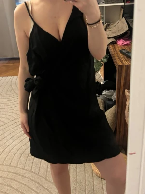 Svart omlottklänning med knyt - Svart kort omlottklänning med smala axelband och knyt i sidan. Klänningen har en v-ringad urringning och är ärmlös, vilket ger en stilren och enkel look. Perfekt för varma dagar eller när du vill känna dig extra snygg.