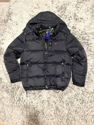 Navy pufferjacka från Polo Ralph Lauren - Snygg navy  pufferjacka från Polo Ralph Lauren med huva och dragkedja framtill. Jackan har två framfickor med dragkedja och den klassiska röda loggan broderad på bröstet. Tillverkad i slitstarkt material, perfekt för kalla vinterdagar.