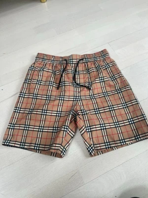Beige rutiga shorts med snörning - Snygga beige shorts med klassiskt rutigt mönster i svart, vitt och rött. Shortsen har elastisk midja med svart snörning och sidofickor. Perfekta för sommaren och ger en trendig vibe till din outfit.