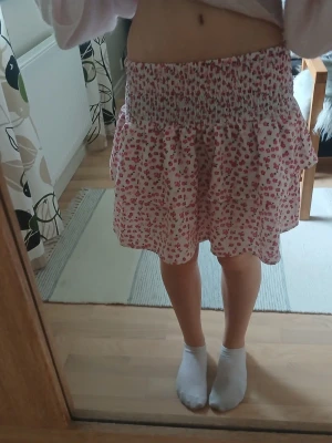 Kort blommig kjol med volang - Söt kort kjol med blommigt mönster i rosa och rött på vit botten. Kjolen har bred smock i midjan och volang nedtill för en luftig och somrig känsla. Perfekt för dig som gillar en feminin och avslappnad stil.