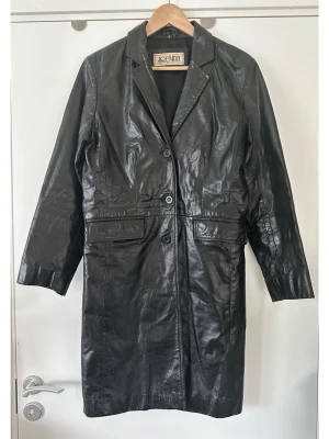 Svart skinnkappa från Jofama, 100% äkta läder - Stilren svart läderkappa / skinnkappa från Jofama i storlek S/36. Kappan har en klassisk design med krage och knäppning framtill. Den har två fickor med lock framtill. En tidlös kvalitetskappa som passar för många tillfällen. 100% äkta läder. Mycket fint skick. 