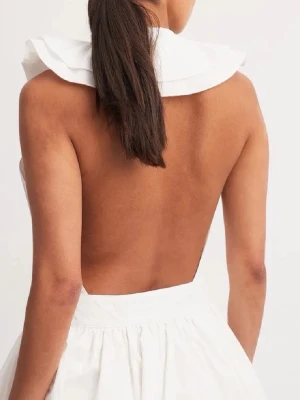 Vit kort klänning med öppen rygg - Säljer en vit klänning med omlottdesign och volangdetaljer framtill. Klänningen är ärmlös och har en öppen rygg som ger en snygg och modern look. Perfekt för sommarens alla tillfällen eller kanske en student! Har en minimal slitning i dragkedjan men ingenting som påverkar användningen eller passformen!