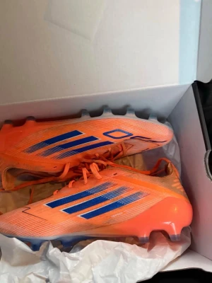 Adidas orange fotbollsskor med blå detaljer - Säljer ett par grymma fotbollsskor från Adidas i en riktigt snygg orange färg med blå ränder och detaljer. Skorna har snörning och är tillverkade i syntetmaterial för bästa prestanda på planen. Medföljer gör en matchande orange påse med blå snören.