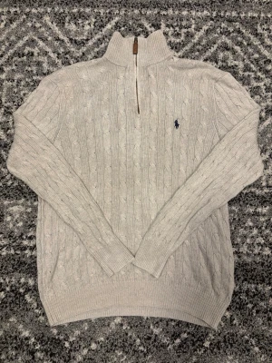 Ljusgrå kabelstickad tröja Polo Ralph Lauren - Ljusgrå kabelstickad tröja från Polo Ralph Lauren med halv dragkedja och hög krage. Tröjan har lång ärm, ribbade muddar och den klassiska mörkblå logotypen broderad på bröstet. Perfekt för lager på lager och en clean look. Tröjan är i storlek M men passar även folk i storleken S. Hör av er om ni har några frågor eller funderingar!!
