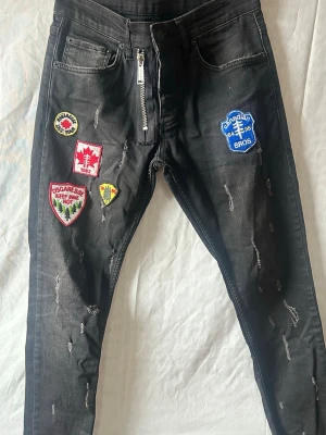 DSQUARED2 varta jeans med patches och dragkedja - Coola svarta jeans med slitningar och flera färgglada patches på framsidan. Jeansen har en unik dragkedja framtill och detaljer som ger en streetwear-vibe. Perfekta för dig som vill sticka ut med din stil.