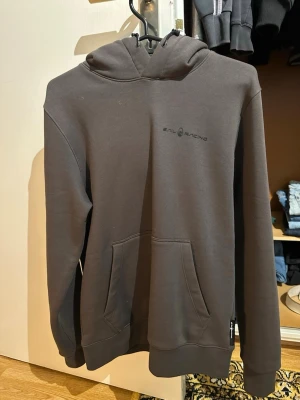 HELT NY Mörkgrå hoodie från Sail Racing - HELT NY Snygg mörkgrå hoodie från Sail Racing med diskret logga på bröstet. Klassisk modell med huva och känguruficka framtill. Mjukt material och ribbade muddar vid ärmslut och nederkant. Perfekt för en avslappnad stil.