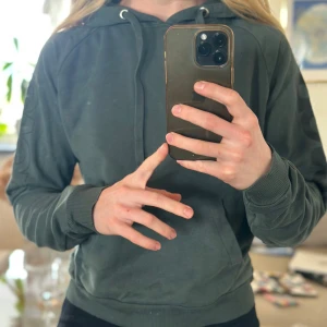 Grön hoodie från Svea - Säljer en snygg grön hoodie från Svea med tryck på armarna. Den har en klassisk design med huva och ribbade muddar. 