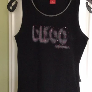 Svart linnetopp från Triumph - Snygg svart linnetopp från Triumph med glittrig text på framsidan. Perfekt för en cool och avslappnad stil. Toppen är ärmlös och har en bekväm passform.