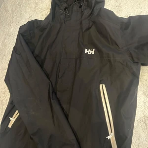 Svart vindjacka från Helly Hansen - Säljer en svart vindjacka från Helly Hansen med dragkedjor i kontrasterande färg. Jackan har en justerbar huva och långa ärmar. Perfekt för blåsiga dagar. Priset går att diskutera vid snabb affär!