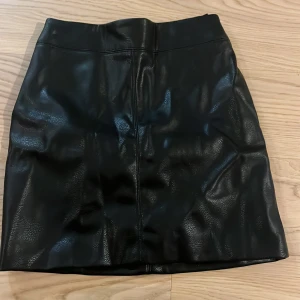 Svart skinnkjol med dragkedja - Snygg svart skinnkjol med dragkedja baktill. Perfekt för en cool och stilren look. Kjolen har en kort längd och en tight passform som framhäver figuren.