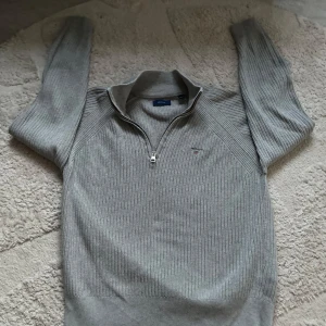 Grå ribbad tröja från GANT - Säljer en stilren grå ribbad tröja från GANT med halv dragkedja och broderad logga på bröstet. Perfekt för kyligare dagar med sin långärmade design och mjuka material. Passar barn i åldern 11-12 år.