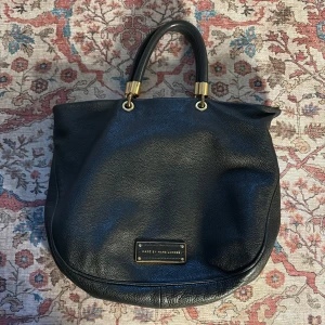 Svart handväska från Marc by Marc Jacobs - Snygg svart handväska i skinn från Marc by Marc Jacobs. Väskan har två handtag med guldfärgade detaljer och en logotypplatta på framsidan. Perfekt för att bära dina dagliga nödvändigheter med stil.