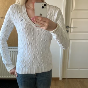 Vit stickad tröja från Ralph Lauren - Säljer en stilren vit stickad tröja från Ralph Lauren med kabelstickat mönster och v-ringning. Tröjan har långa ärmar och en liten broderad logga på bröstet. nypris: runt 1000 kr