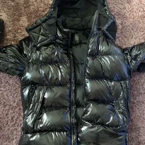 Svart glansig pufferjacka från Boss - Snygg svart pufferjacka från Boss med glansig finish. Jackan har en dragkedja framtill och är perfekt för kyligare dagar. Den har en klassisk design med långa ärmar och en hög krage för extra värme.Jag har haft den i 1år och den är inte sönder eller nått sånt 