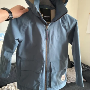 Blå regnjacka från Tretorn - Säljer en snygg blå regnjacka från Tretorn med huva och dragkedja framtill. Jackan har två praktiska fickor och är perfekt för regniga dagar. Den är långärmad och har en stilren design.