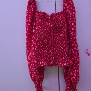 Röd blommig blus - Charmig röd blus med vita blommor och långa ärmar. Blusen har en smockad design med knytning framtill och volangdetaljer vid fållen. Perfekt för en romantisk look.