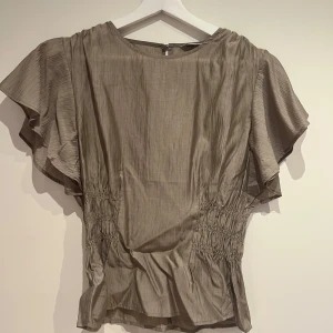 Beige blus med korta ärmar - Säljer en elegant beige blus med korta ärmar och rynkade detaljer i midjan. Blusen har en rund hals och en diskret knäppning i ryggen. Perfekt för en stilren look.