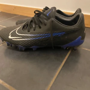 Svarta fotbollsskor från Nike - Säljer ett par svarta Nike PhantomX fotbollsskor med blå detaljer och snörning. Skorna har en robust sula med dobbar, perfekt för gräsplaner. Den stilrena designen kombinerar funktionalitet med en sportig look. Köptes för 1000 kr och används bara i en månad. 