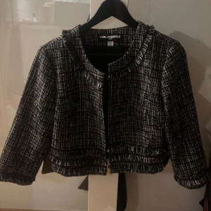 Svartvit kavaj från Karl Lagerfeld - Elegant svartvit kavaj från Karl Lagerfeld med ett klassiskt rutigt mönster. Kavajen har en chic, croppad design med dekorativa fransdetaljer vid kragen och nederkanten. Perfekt för en stilfull look.