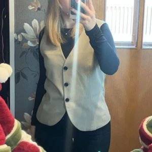 Beige väst med knappar - Snygg beige väst med knappar framtill. Perfekt för att lägga till en stilren touch till din outfit. Passar bra över en långärmad tröja.