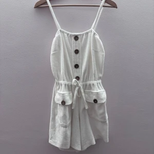 Vit ärmlös jumpsuit med knappar - Säljer en söt vit ärmlös jumpsuit med knappar framtill och knytband i midjan. Den har praktiska fickor med knappar och smala axelband. Perfekt för varma dagar! är också i ett handuks liknande material 