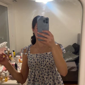 Blommig ärmlös topp med knytband - Säljer en söt blommig topp med blå blommor på vit bakgrund. Toppen är ärmlös och har knytband på axlarna för en justerbar passform. Perfekt för varma sommardagar!