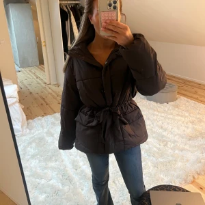 Trendig vinterjacka  - Säljer en trendig brun dunjacka med bälte i midjan. Jackan har långa ärmar och dragkedja framtill. Perfekt för kyliga dagar och ger en snygg siluett. 