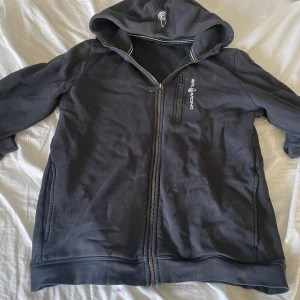 Svart  hoodie från Sail Racing - Säljer en mörkgrå hoodie från Sail Racing med dragkedja och huva. Den har en broderad logga på bröstet och ribbade muddar. Perfekt för en avslappnad stil.