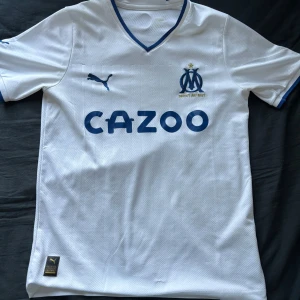 Marseille Hemma 22-23 - Säljer min Marseille tröja från säsongen 22/23 från Puma med blå detaljer. Tröjan har korta ärmar och en V-ringad krage. Den har en logotyp på bröstet och texten 'CAZOO' i blått. Perfekt för fotbollsfans!